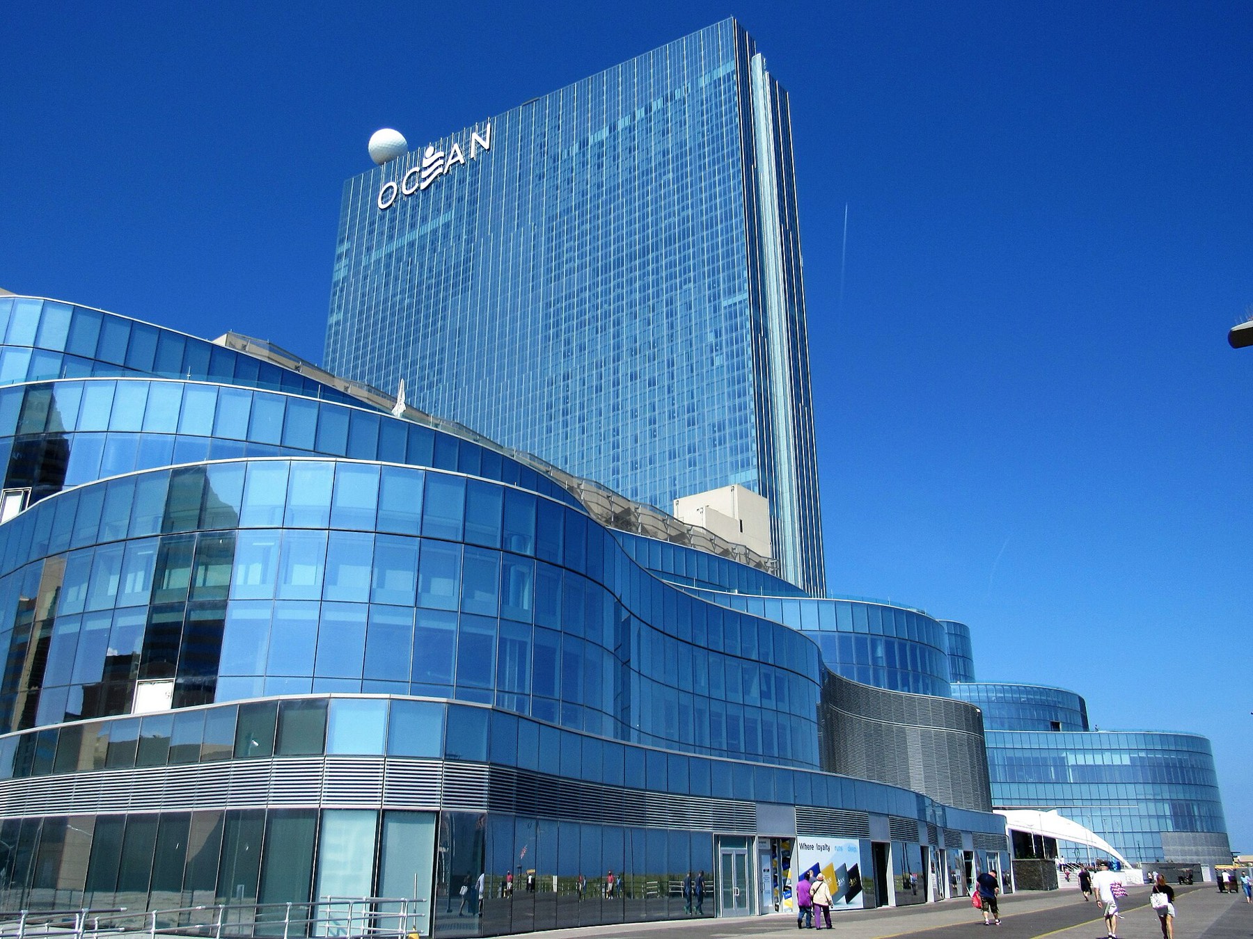 The curved blue-glass podium and tall glass tower of Ocean Casino Resort on the Atlantic City Boardwalk under a clear sky