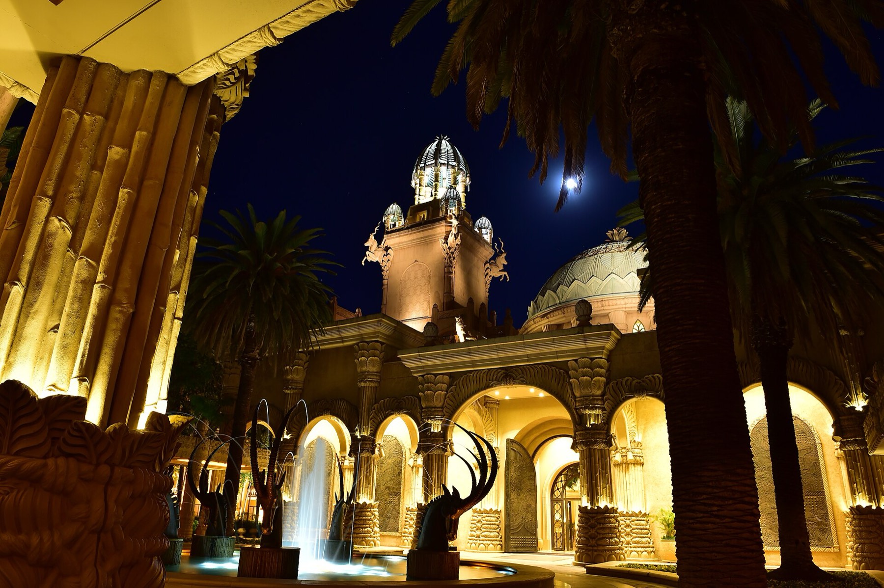 The floodlit arched entrance court of the Palace of the Lost City at night, with domed towers, palms and bronze sable antelope fountains